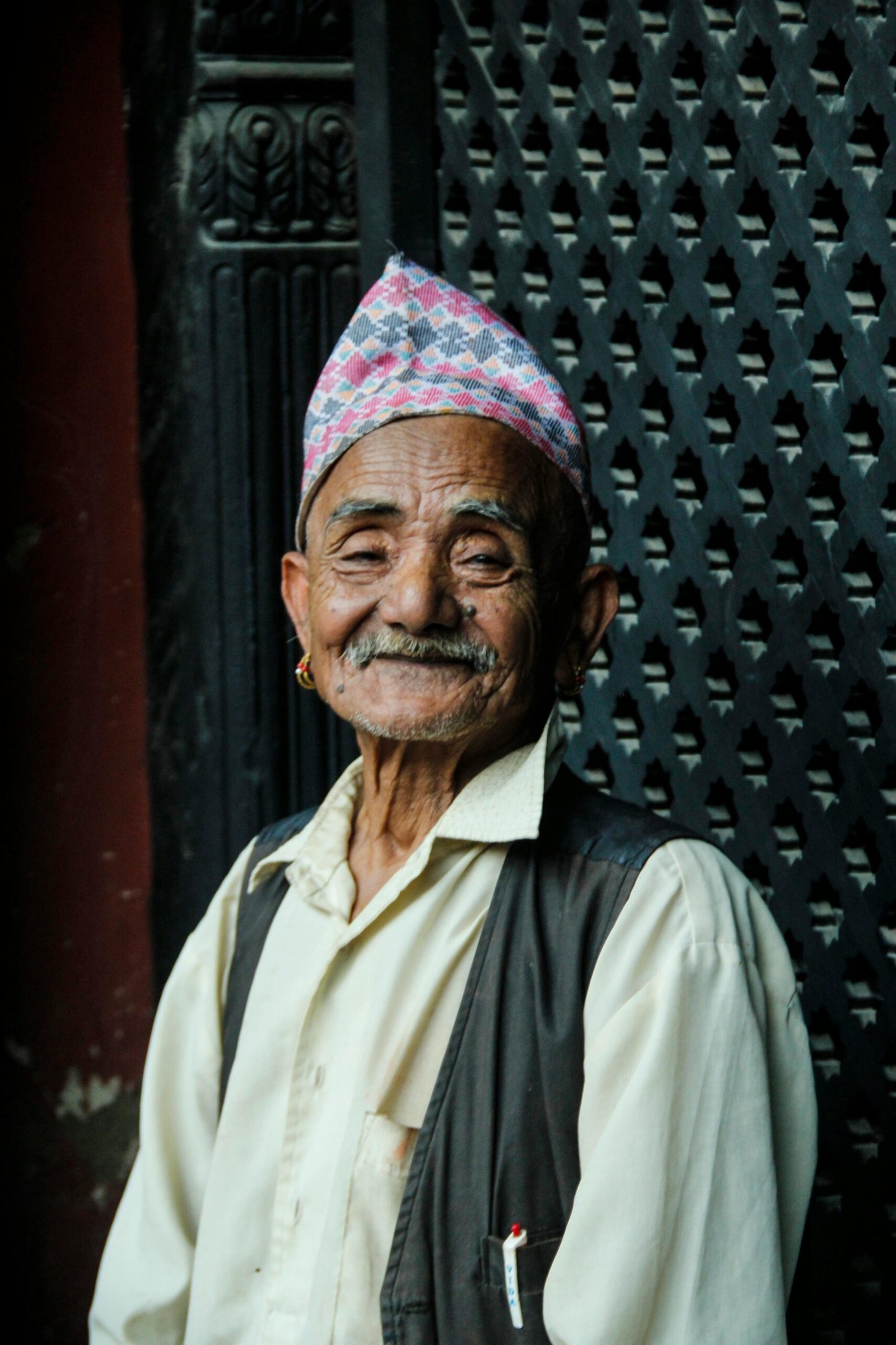 A portrait of an elderly Nepali man wearing traditional attire, smiling outdoors in Patan.